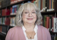 Janis Winn in glasses, white shirt and pink blazer stands in front of library stacks.