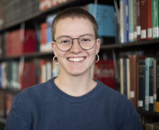 Glory Stevens in blue shirt and wearing glasses stands in front of library stacks.