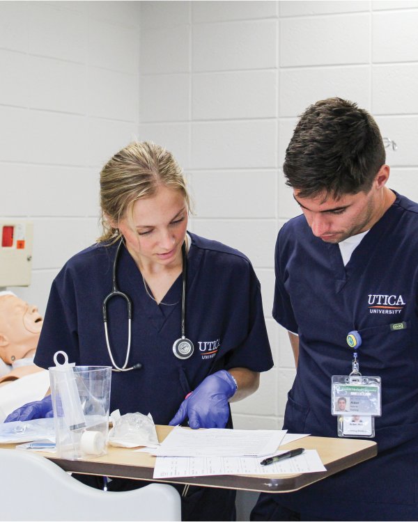 Nursing students in scrubs, look over paperwork near a mock-patient.