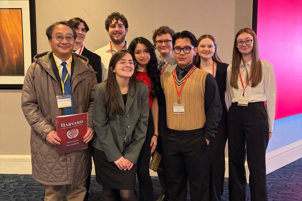 Students stand with professor, smiling, while holding a folder that says "Harvard" on it.