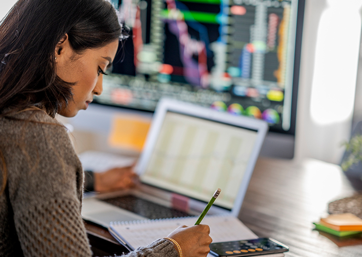 woman writing calculations on paper in front of chart on screen